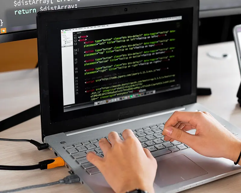 Manos escribiendo código en el teclado de un portátil sobre un escritorio de oficina, con un monitor de código detrás